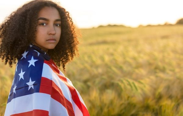 Sad depressed mixed race African American girl teenager female young woman with tears in her eyes in a field of wheat or barley crops holding and wrapped in USA stars and stripes flag in golden sunset evening sunshine
